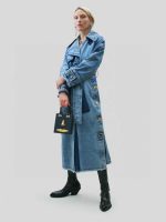 A full-length shot of a stylish women's denim trench coat. The coat is long with a belted waist, large gold buttons down the front, and frayed seams at the bottom.