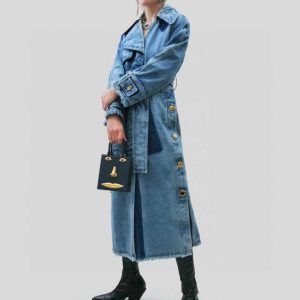 A full-length shot of a stylish women's denim trench coat. The coat is long with a belted waist, large gold buttons down the front, and frayed seams at the bottom.