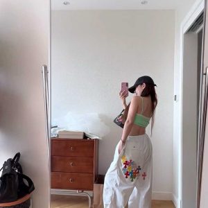 Person wearing white Chrome Hearts style cargo pants with colorful cross patchwork on the left leg, a light green top, and a black baseball cap